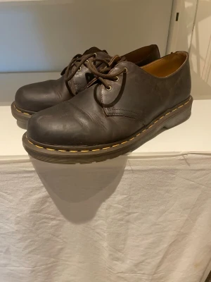 Bruna Dr. Martens loafers i läder - Säljer ett par klassiska bruna loafers från Dr. Martens i äkta läder. Skorna har rund tå, snörning och den ikoniska gula sömmen runt sulan. Perfekta för dig som gillar en tidlös och robust stil.