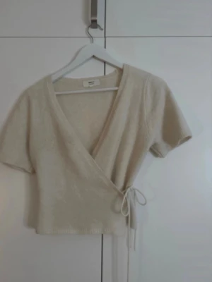 Beige omlottopp med knyt - Snygg beige omlottopp med korta ärmar och knyt i sidan. Toppen har v-ringad hals och är croppad, vilket ger en trendig look. Perfekt att matcha med höga jeans eller kjol för en chill stil.