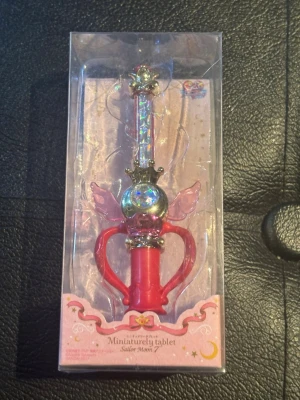 Sailor Moon miniatyr trollstav - Magisk miniatyr trollstav från Sailor Moon med skimrande detaljer, rosa handtag och vingar samt guldfärgade inslag. Perfekt accessoar för cosplay eller maskerad, levereras i originalförpackning med genomskinligt lock.