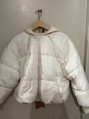 Vit pufferjacka med huva - Säljer en oversized vit pufferjacka med dragkedja och huva. Jackan har quiltad design och breda kanaler som ger en riktigt fluffig look. Perfekt för kalla dagar när du vill hålla dig varm och samtidigt se trendig ut.