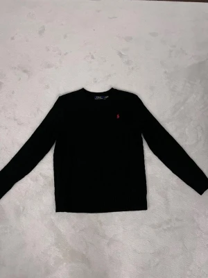 Svart kabelstickad tröja Polo Ralph Lauren - Svart kabelstickad tröja från Polo Ralph Lauren med rund halsringning och klassisk röd broderad logga på bröstet. Tröjan har långa ärmar och ribbade muddar vid ärmslut och nederkant. Perfekt för en stilren look.