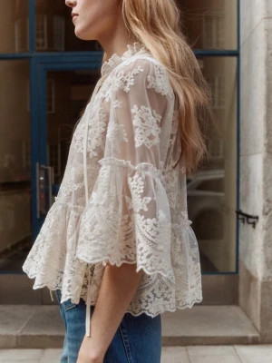 Vit spetsblus med volang och knytband - Superfin transparent vit blus i spets med blommigt mönster, volang nertill och knytband vid halsen. Blusen har trekvartsärm och romantisk känsla, perfekt att styla lager på lager. Materialet är tunn mesh med broderade detaljer.