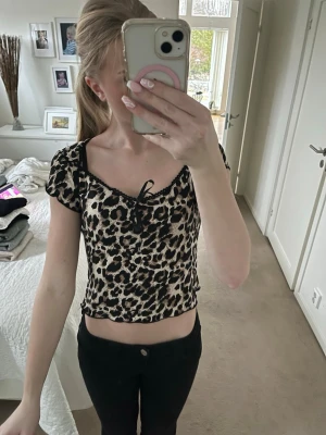 Leopardmönstrad croppad topp med snörning - Säljer en leopardmönstrad croppad topp med korta ärmar och snörning framtill. Toppen har en v-ringad design med små volangdetaljer längs halsen. Perfekt för dig som gillar trendiga och djurmönstrade plagg.