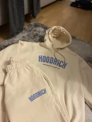 Beige Hoodrich loungewear set - Snyggt beige loungewear set från Hoodrich med matchande hoodie och byxor. Setet har blå logotyptryck på både tröjan och byxorna. Hoodien har huva och dragsko, byxorna har resår i midjan för extra komfort. Perfekt för chill dagar hemma.