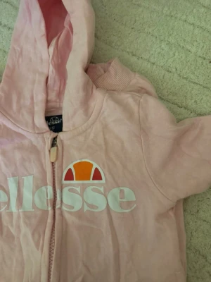Ljusrosa Ellesse overall med dragkedja - Snygg ljusrosa mjukis overall från Ellesse med vit logga och dragkedja framtill. Overallen har huva och ribbade muddar vid ärmsluten. 