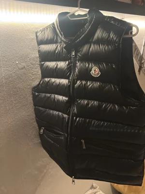 Moncler gui - Snygg svart dunväst från Moncler med hög krage och två dragkedjefickor framtill. Västen har Moncler-logga på bröstet och quiltad design som ger en modern look. Perfekt att slänga över en hoodie eller tröja för extra värme.