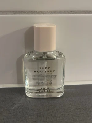 Zara Nude Bouquet parfym - Fräsch parfym från Zara med namnet Nude Bouquet. Flaskan är genomskinlig med en ljusbeige, nästan nude-färgad kork och stilren design. Glasflaskan har raka linjer och minimalistisk text framtill. Perfekt för dig som gillar eleganta och moderna dofter.