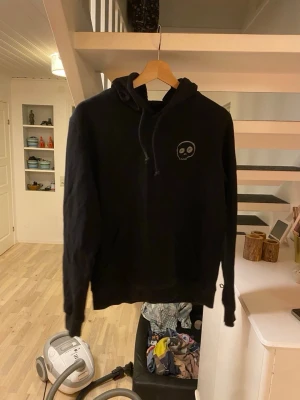Svart hoodie med döskalletryck - Säljer en svart hoodie med huva och dragsnören. På bröstet finns ett litet döskalletryck och på ryggen ett större tryck med en tecknad skelettfigur och texten 'Who? What?'. Perfekt för dig som gillar streetwear och vill sticka ut lite extra.