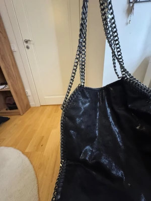 Svart axelväska med kedjedetaljer - Cool svart axelväska med glansig finish och kraftiga silverfärgade kedjor som axelrem och runt kanterna. Väskan har en mjuk, slouchy form och ger en edgy vibe till din outfit. Perfekt för dig som gillar statement-accessoarer.