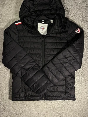 Svart dunjacka från Rossignol - Snygg svart dunjacka från Rossignol med huva och dragkedja framtill. Jackan har quiltad design, patch med logga på ärmen och coola röd-vita detaljer på axeln. Perfekt för kalla vinterdagar och riktigt stilren look.