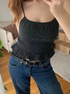 Svart smockad linnetopp med volang - Trendig mörkblå linnetopp med smala axelband och smockad struktur. Toppen har en lätt volangkant nedtill och sitter figurnära. Perfekt till jeans eller kjol för en enkel men snygg look.