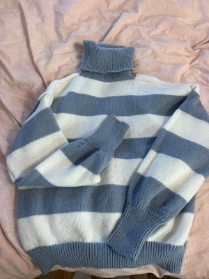 Randig polotröja i blått och vitt - Stickad polotröja med breda blå och vita ränder. Tröjan har långa ärmar, ribbstickade muddar och hög krage. Tillverkad i 100% polyester för en mjuk och bekväm känsla. Perfekt för kyliga dagar. Stl M men skulle säga att det är storlek S