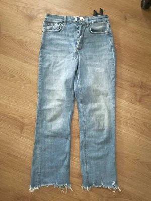 Ljusblå raka jeans med rå kant - Säljer ett par ljusblå jeans med raka ben och trendig rå kant nertill. Jeansen har knäppning med flera knappar framtill och klassiska fem fickor. Materialet är denim i bomull och passformen är avslappnad och lite croppad.