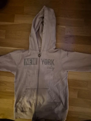Beige hoodie med New York-tryck - Säljer en rosa hoodie med dragkedja och huva. På bröstet står det 'NEW YORK city' i grått tryck. Hoodien har två fickor framtill och ribbade muddar vid ärmar och nederkant. Perfekt för en avslappnad streetstyle-look.