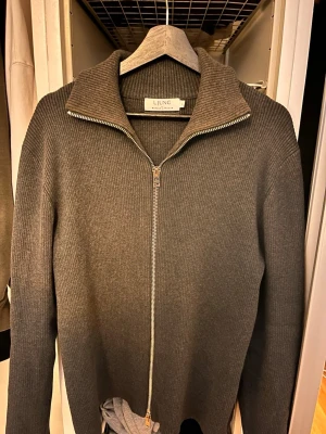 Grå ribbad ziptröja från Ljung - Snygg grå ribbstickad tröja från Ljung med hel dragkedja framtill och bred krage. Tröjan har lång ärm och är tillverkad i ett mjukt, tjockare material som passar perfekt när det är kyligt ute. Enkel och stilren design som funkar till det mesta. Storlek L men sitter som M. Mörkgrå