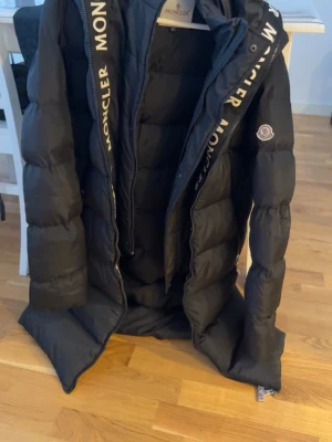 Svart Moncler dunjacka med logga - Snygg svart dunjacka från Moncler med stor logotyp på dragkedjan och klassisk Moncler-patch på ärmen. Jackan har en puffig design, hög krage och dragkedja framtill. Perfekt för kalla vinterdagar och riktigt trendig stil.