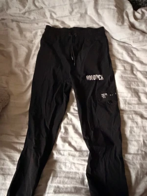 Svarta mjukisbyxor från Hoodrich - Svarta mjukisbyxor från Hoodrich med tryck på ena benet och snörning i midjan. Byxorna har smal passform och resår i midjan. Materialet ser ut att vara mjukt och bekvämt, perfekt för chill dagar. Snygg streetstil med vita detaljer och texttryck.