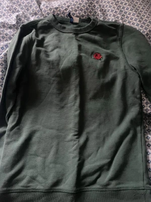 Grön sweatshirt med rosbrodyr - Mysig grön sweatshirt med rund hals och en liten röd ros broderad på bröstet. Tröjan har ribbade muddar vid ärmslut och nederkant. Perfekt för en chill och stilren look.