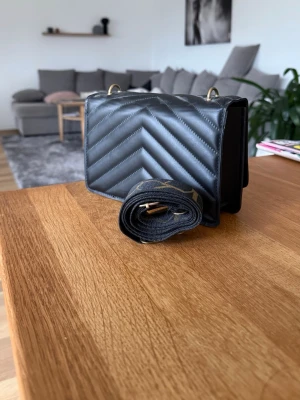 Svart quiltad handväska med axelrem - Snygg svart handväska i quiltat mönster med chevron-detaljer. Väskan har guldfärgade metalldetaljer och en bred avtagbar axelrem. Perfekt storlek för det viktigaste och ger en trendig look till din outfit.