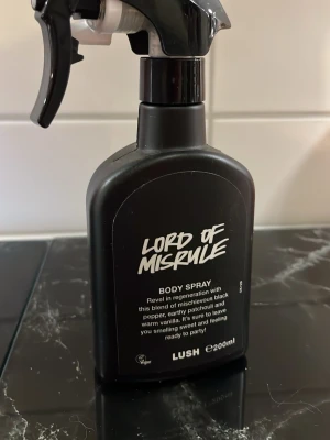 Lush Lord of Misrule Body Spray - Lord of Misrule Body Spray från Lush i svart plastflaska med spraymunstycke. Doften har inslag av svartpeppar, patchouli och varm vanilj. Flaskan rymmer 200 ml och har en modern, edgy design med vit text på svart bakgrund.