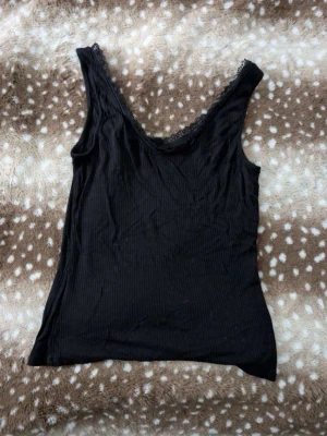 Svart ribbat linne med spetskant - Svart linne i ribbat material med djup v-ringning och fin spetskant längs halsringningen. Perfekt att styla lager på lager eller bära som det är. Enkel och stilren design som passar till mycket.