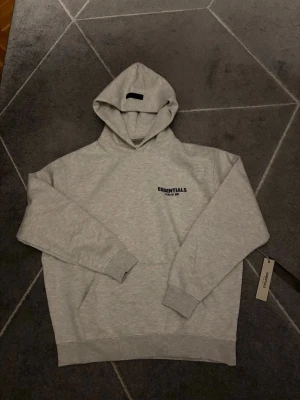Grå Essentials Fear of God hoodie - Snygg ljusgrå hoodie från Essentials Fear of God med svart logga på bröstet och stor text på ryggen. Klassisk passform med huva och ribbade muddar. Perfekt för en avslappnad och trendig streetwear-look.
