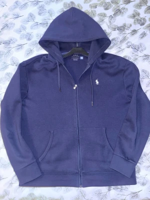 Mörkblå hoodie från Polo Ralph Lauren - Snygg mörkblå hoodie från Polo Ralph Lauren med dragkedja och klassisk logga broderad på bröstet. Tröjan har huva med snören, två fickor framtill och ribbade muddar. Perfekt för en avslappnad och cool look.