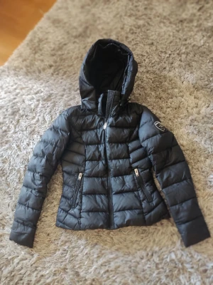 Svart dunjacka Rockandblue dam - Snygg svart dunjacka från Rockandblue med huva och två dragkedjeförsedda fickor. Jackan har quiltad design och en patch på ena ärmen. Perfekt för kalla vinterdagar och riktigt skön att bära.