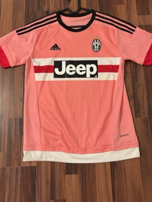 Juventus borta tröja 2015/16 ( Pogba ) - Juventus rosa matchtröja från Adidas med Pogba och nummer 6 på ryggen. Tröjan har svarta detaljer, röda ärmslut och Jeep-logga på bröstet. Tillverkad i lätt och ventilerande polyester, perfekt för fotbollsträning eller match.