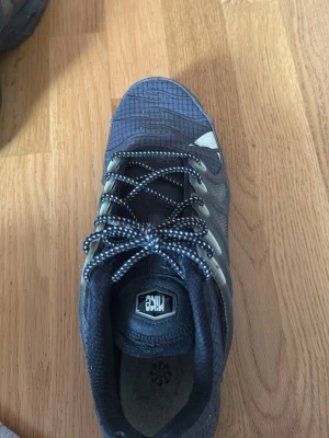 Nike svarta sneakers med mönstrad sula - Nike sneakers i svart med grå detaljer och reflekterande partier. Skorna har en mönstrad sula och svarta/vita skosnören. Ovandelen är i textil med struktur och plastdetaljer på sidorna. Märket syns tydligt på plösen och hälen.