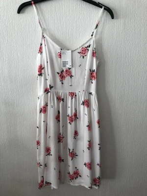 Vit blommig kortklänning från H&M - Söt vit kortklänning från H&M med rosa blommigt mönster och smala axelband. Klänningen har en smockad rygg och lätt v-ringad front, vilket ger en bekväm och luftig passform. Perfekt för varma dagar och somriga vibes. Superfin och superskön tyg. 