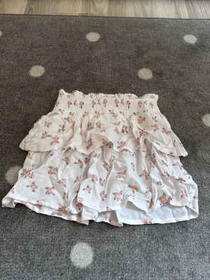 Vit blommig volangkjol - Supersöt kort kjol i vitt med små rosa blommor och volangdetaljer. Kjolen har en smockad midja som ger en bekväm passform och ett romantiskt uttryck. Perfekt för dig som gillar en feminin och lekfull stil.