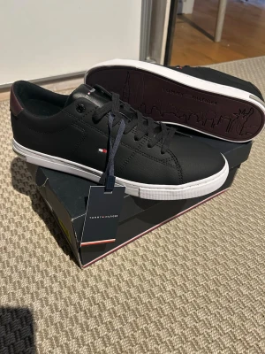 Tommy Hilfiger Skor - Skorna är helt nya men däremot så är självaste sko kartongen lite trasig. De är svarta med en vit rem och på baksidan är den en vinröd flärp och vid sko sulan är det en grå plats bit där de står ”Tommy himfiger”