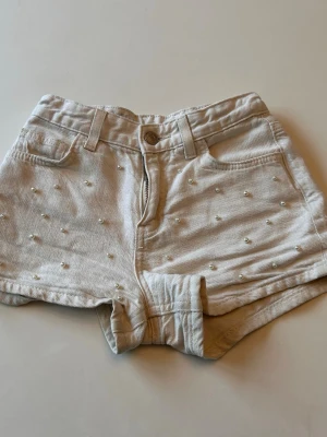 Beige shorts med pärlor - Snygga beige shorts med pärldetaljer framtill. Klassisk femficksmodell med bälteshällor och knappstängning. Perfekta för sommaren och ger en trendig touch till din outfit.