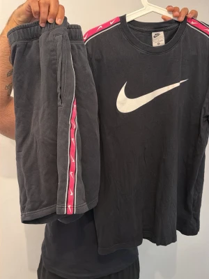 Nike svart set med rosa detaljer - Snyggt set från Nike med långärmad svart tröja och matchande shorts. Båda plaggen har vita Nike-loggor och coola rosa detaljer längs axlar och sidor. Tillverkade i mjukt bomullsmaterial för en bekväm känsla. Perfekt för dig som gillar sportig och avslappnad stil.