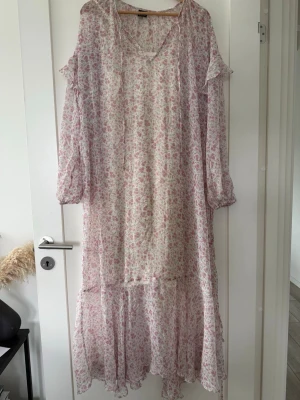 Blommig långklänning från Gina Tricot - Långklänning från Gina Tricot i vitt med rosa och grönt blommönster. Klänningen har volangdetaljer, v-ringning med knytband och långa ärmar med resår vid handlederna. Luftig och flowy passform, perfekt för varma dagar.