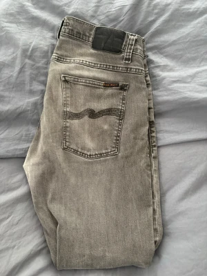 Grå jeans från Nudie Jeans - Snygga grå jeans från Nudie Jeans med klassisk femficksdesign och vågig detalj på bakfickan. Jeansen har en rak passform och är tillverkade i mjukt denimtyg. Perfekta för dig som gillar stilrena och tidlösa jeans.