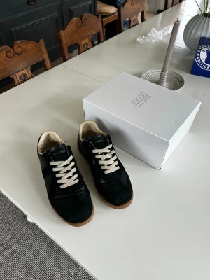  Maison Margiela Gats Svarta - Maison Margiela sneakers i svart läder och mocka med vita skosnören och beige innersula. Klassisk låg modell med gummisula och stilren design. Perfekta för dig som gillar minimalistisk och tidlös stil.