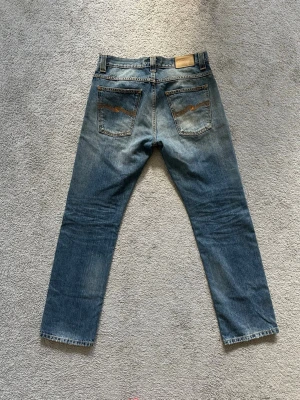 Nudie Jeans Avergage Joe - Säljer ett par klassiska blå raka jeans från Nudie Jeans i modellen Average Joe, med snygga slitningar och kontrastsömmar. Jeansen har fem fickor, knappgylf och ikoniska broderade bakfickor. Storlek: W31, L32. Midja: 41 cm. Ytterbenslängd: 104 cm.
