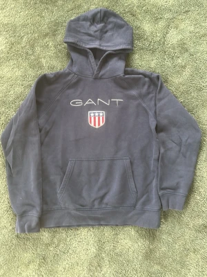 Mörkblå hoodie från GANT - Mörkblå hoodie från GANT med klassisk logotyp och ett broderat emblem på bröstet. Tröjan har en stor huva och en praktisk magficka. Materialet är mjukt och bekvämt, perfekt för en avslappnad stil. Snygg design med ribbade muddar och rak passform.