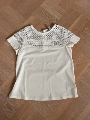 Vit blus med spetsdetaljer H&M - Säljer en söt vit blus från H&M i storlek XS. Toppen har korta ärmar och en övre del i broderad spets med hålmönster som ger en fin vibe. Perfekt till jeans eller kjol för en fräsch look.