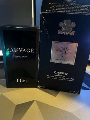 Dior Sauvage & Creed Aventus parfym - Två exklusiva parfymer: Dior Sauvage Eau de Parfum i svart, stilren flaska med vit text och Creed Aventus i svart kartong med silverdetaljer och klassisk logga. Båda är populära dofter för den som vill sticka ut med elegans.