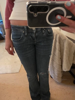 Blå slitna bootcut jeans - Säljer ett par blå bootcut jeans med slitningar vid knäna. Jeansen har klassisk femficksmodell, låg midja och är tillverkade i denim. Perfekta för en avslappnad och trendig look.