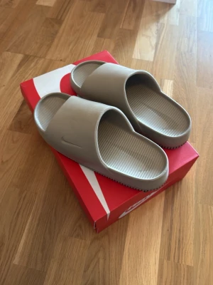 Beige slides från Nike - Snygga beigea tofflor från Nike med minimalistisk design och diskret logga på sidan. Tillverkade i ett mjukt och flexibelt syntetmaterial med räfflad innersula för extra komfort. Perfekta för att chilla hemma eller dra till stranden.