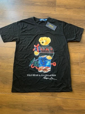 Svart Polo Bear t-shirt från Ralph Lauren - Svart t-shirt från Ralph Lauren med Polo Bear-tryck framtill. Björnen har en julig tröja och håller ett paket, med texten 'POLO BEAR by RALPH LAUREN' under motivet. Klassisk rund hals och korta ärmar. Perfekt statement-plagg för dig som gillar ikoniska prints.