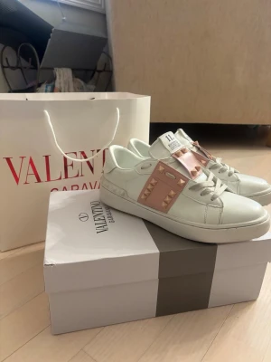Valentino Garavani vita sneakers - Snygga vita sneakers med en bred rosa rem över foten med nitar. Skorna har en klassisk låg siluett och är tillverkade i läder. Perfekta för dig som vill ha en exklusiv och trendig look. Skorna har några defekter, en skada framtill på ena foten och en nit har åkt av men det är inget som syns så mycket. 