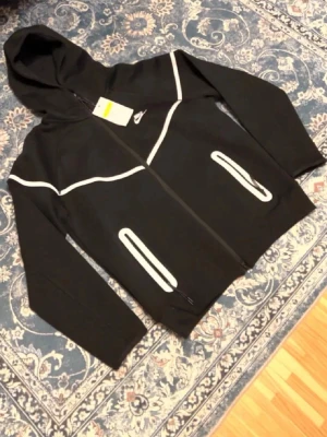 Svart Nike hoodie med reflexdetaljer - Svart hoodie från Nike med dragkedja och vita reflexdetaljer längs bröstet och vid fickorna. Tröjan har huva och två fickor med dragkedja. Perfekt för dig som vill sticka ut med coola detaljer och samtidigt synas i mörkret. Materialet ser ut att vara mjukt och bekvämt.