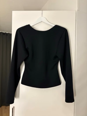 Svart  långärmad topp - Stilren svart topp med lång ärm och v-ringning baktill. Toppen har en figurnära passform och är tillverkad i ett slätt, stretchigt material. Perfekt för dig som gillar minimalistisk och modern stil.