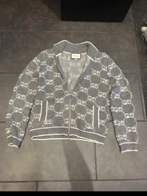 Grå ziptröja med GG-mönster från Gucci - Snygg grå ziptröja från Gucci med ikoniskt GG-mönster i vitt över hela plagget. Tröjan har ribbade muddar, krage och nederkant samt vita detaljer vid fickorna. Perfekt för dig som vill ha en exklusiv och trendig look.