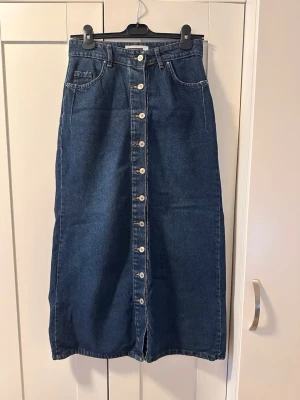 Lång jeanskjol med knappar - Snygg lång jeanskjol i mörkblå denim med raka ben och knappar hela vägen framtill. Klassisk femficksmodell och hög midja. Perfekt till vardags för en avslappnad men trendig look.